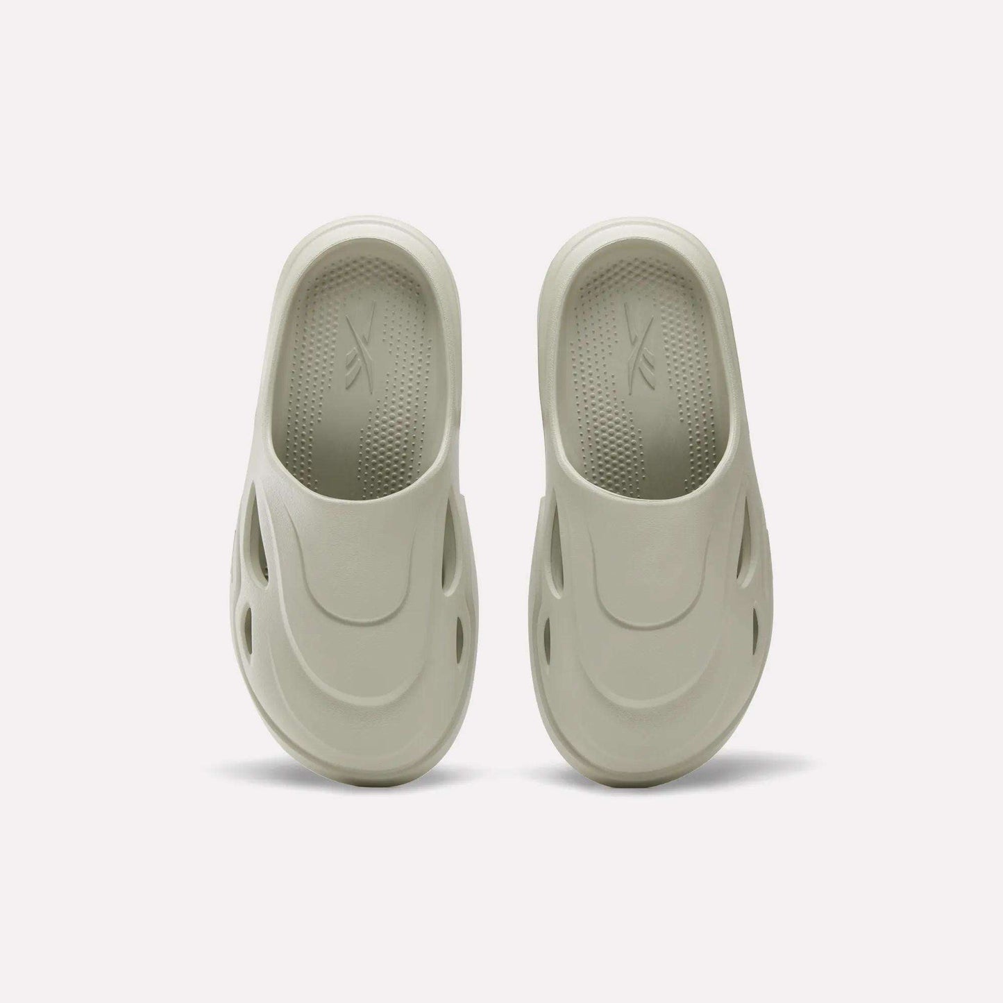 The Reebok DMX Recovery Mules in light beige are shown on a plain white background, featuring molded EVA design, rounded toes, side ventilation holes, and textured insoles inspired by DMX Moving Air tech.