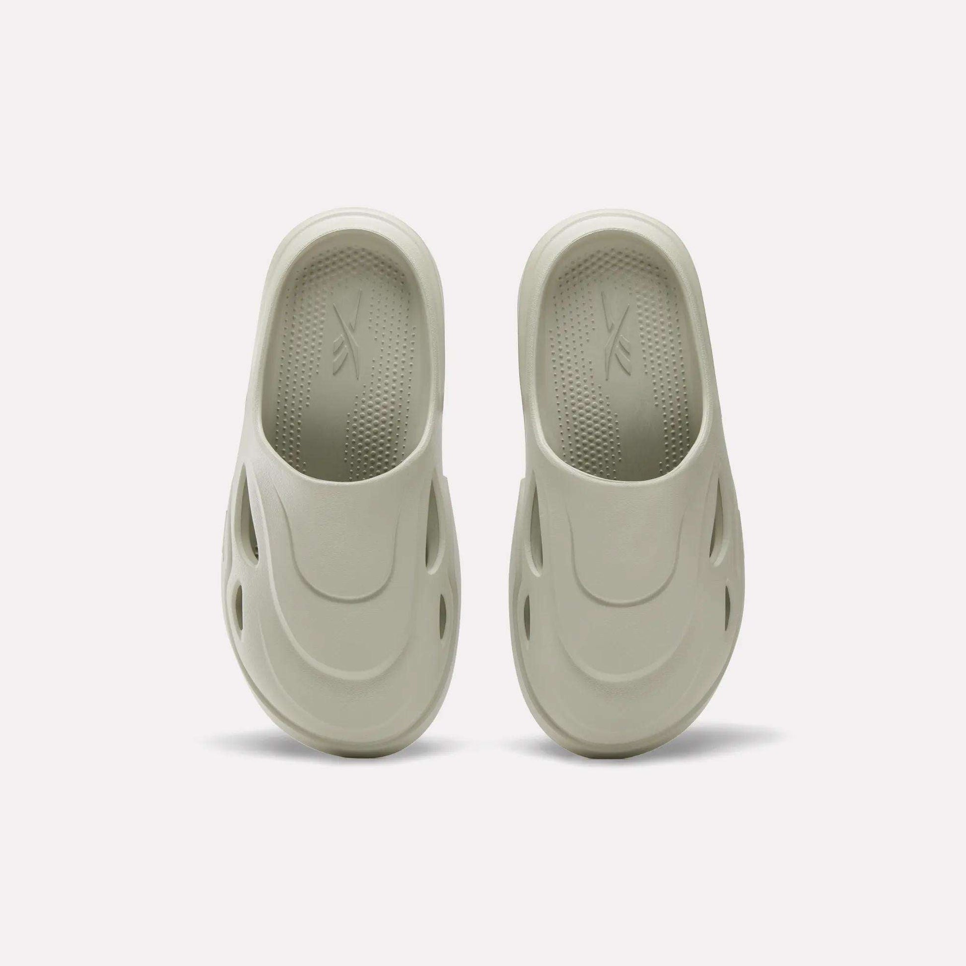 The Reebok DMX Recovery Mules in light beige are shown on a plain white background, featuring molded EVA design, rounded toes, side ventilation holes, and textured insoles inspired by DMX Moving Air tech.