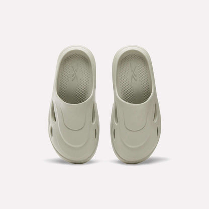 The Reebok DMX Recovery Mules in light beige are shown on a plain white background, featuring molded EVA design, rounded toes, side ventilation holes, and textured insoles inspired by DMX Moving Air tech.
