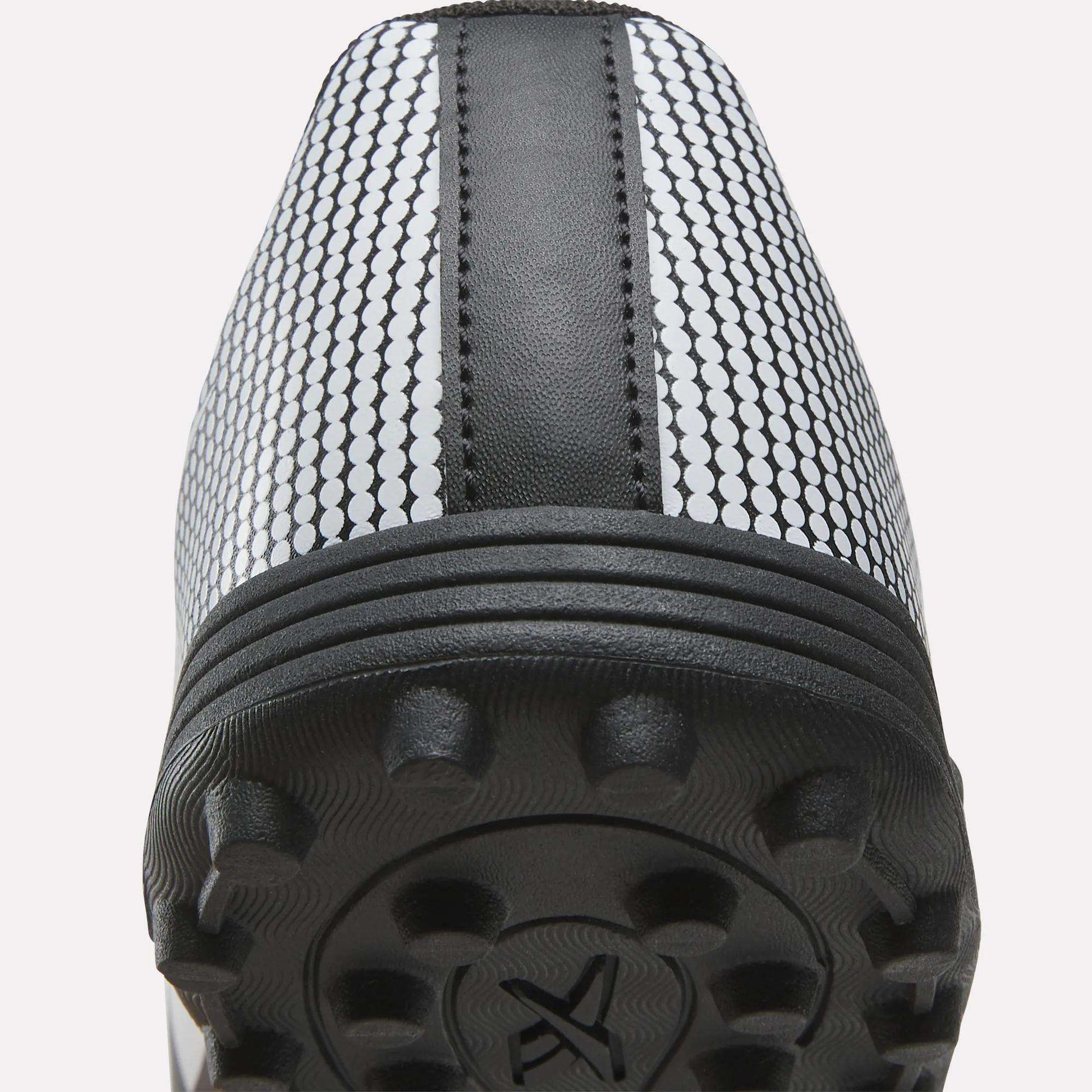 Close-up view of the heel and sole of the Reebok Strike Turf Soccer Shoes, showcasing textured rubber studs, a central black stripe, and a white upper patterned with small black circles—ideal as practice soccer shoes.
