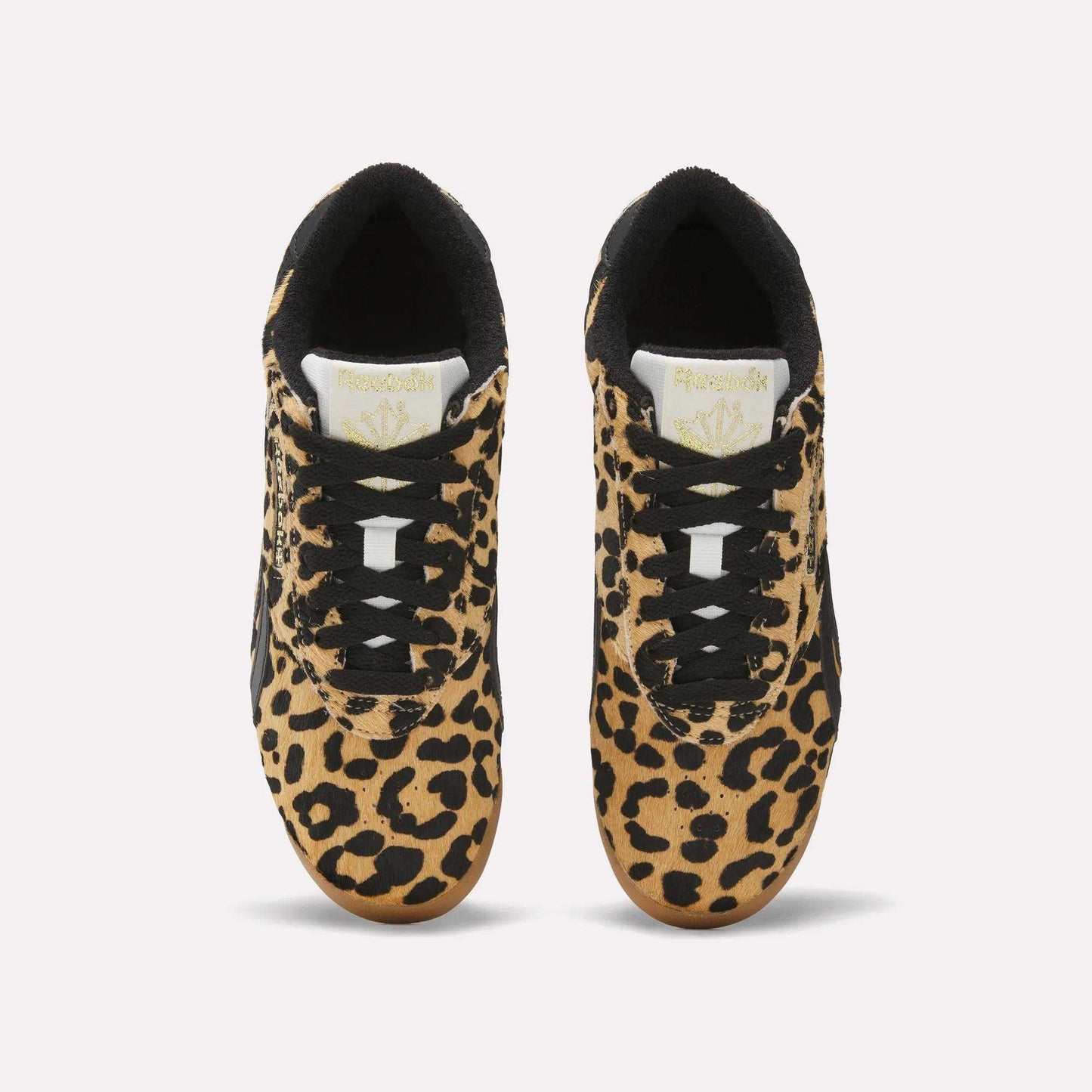A pair of Reebok Women's Freestyle Lo Shoes with a bold leopard print pony hair upper, black laces and accents, and white tongues featuring the gold Reebok logo, inspired by classic '80s fitness trainers, viewed from above on a white background.