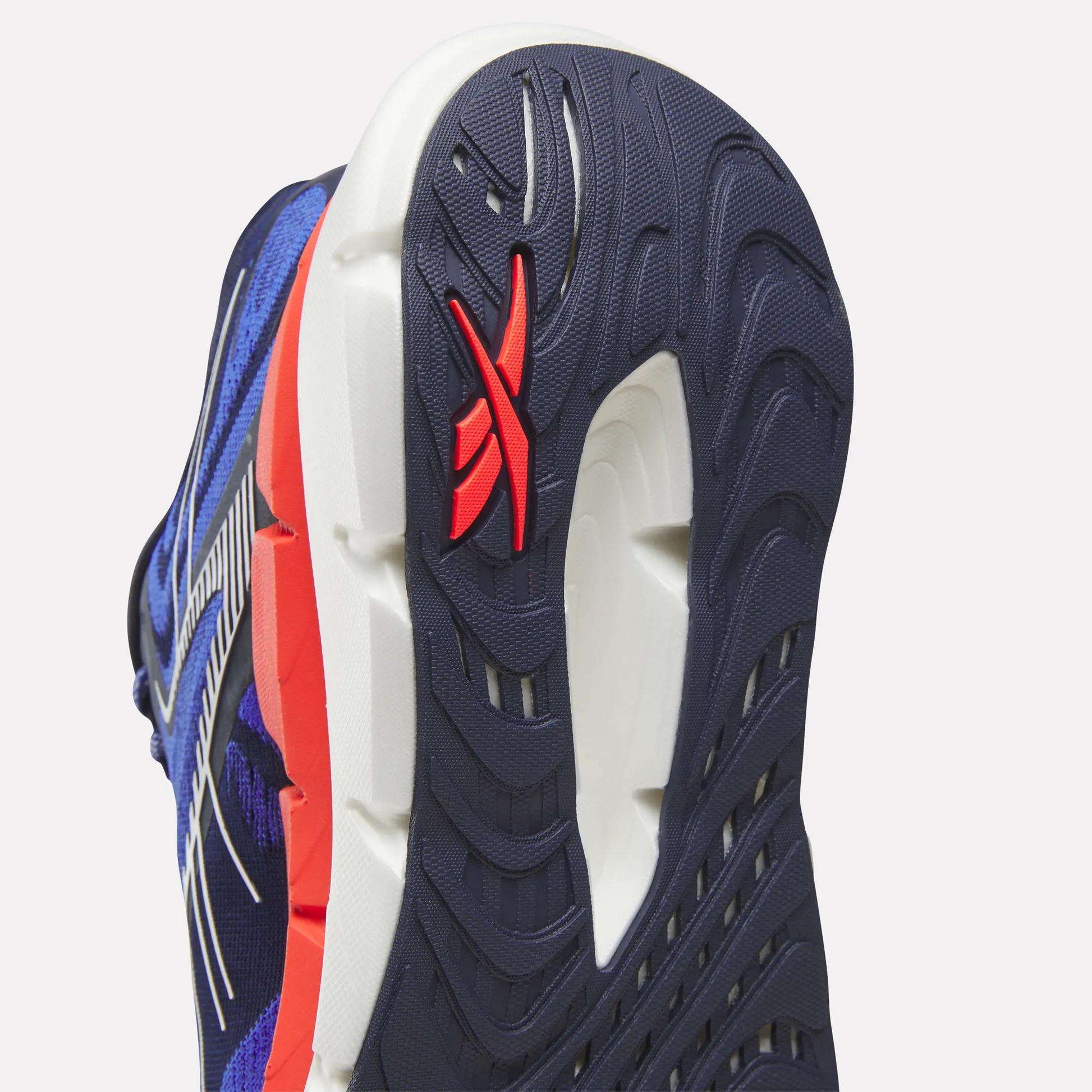 Close-up of the sole on Reebok Men's FloatZig Double Running Shoes, featuring a blue, white, and red color scheme. The outsole has textured grooves and a red Reebok logo on the heel for max-cushioned running comfort.