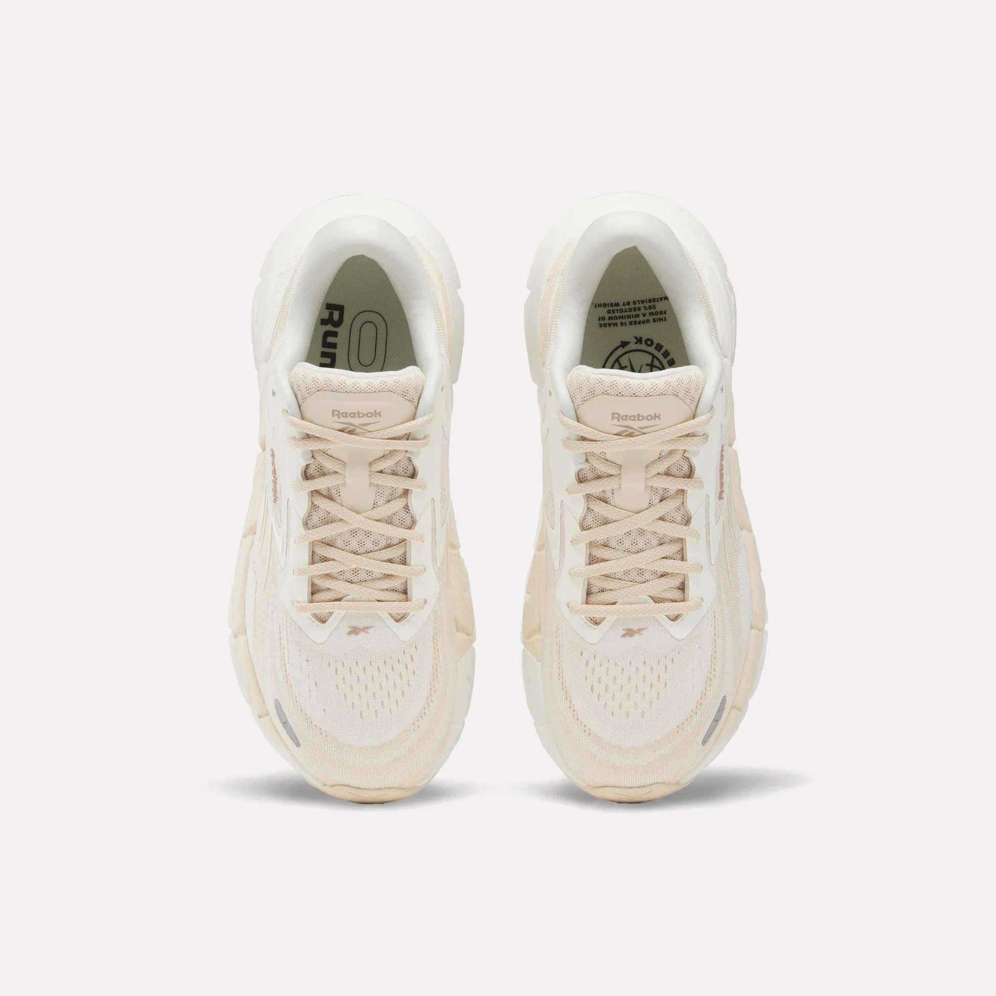 A pair of Women's FloatZig Double Running Shoes by Reebok in beige are shown from above, featuring laces, mesh texture, and logo details on the tongues—designed for max-cushioned runs—set against a plain white background.
