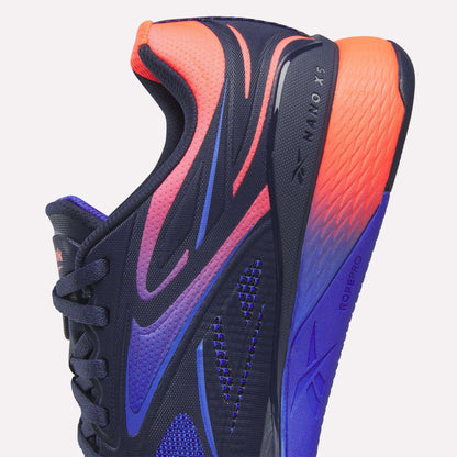 Close-up of Reebok Nano X5 Edge Training Shoes in navy blue with bold purple, pink, and orange gradients. Features textured mesh, black laces, and “Nano X5 Edge” with “RopePro” visible on the side and sole for superior performance.