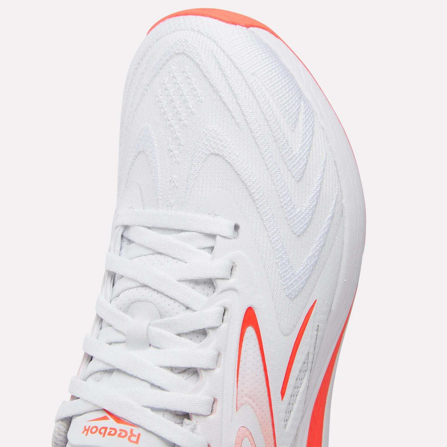 Close-up of the Reebok Women's Nano X5 Edge Training Shoes in white with orange accents, showcasing a wavy-patterned upper, white laces, and a partially visible logo near the toe. Perfect addition to any women’s HIIT shoe collection.