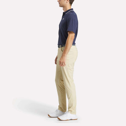 A man in profile wears the Reebok Men's BDC Signature Polo Shirt, beige pants, a white belt, and white shoes, standing against a plain white background.