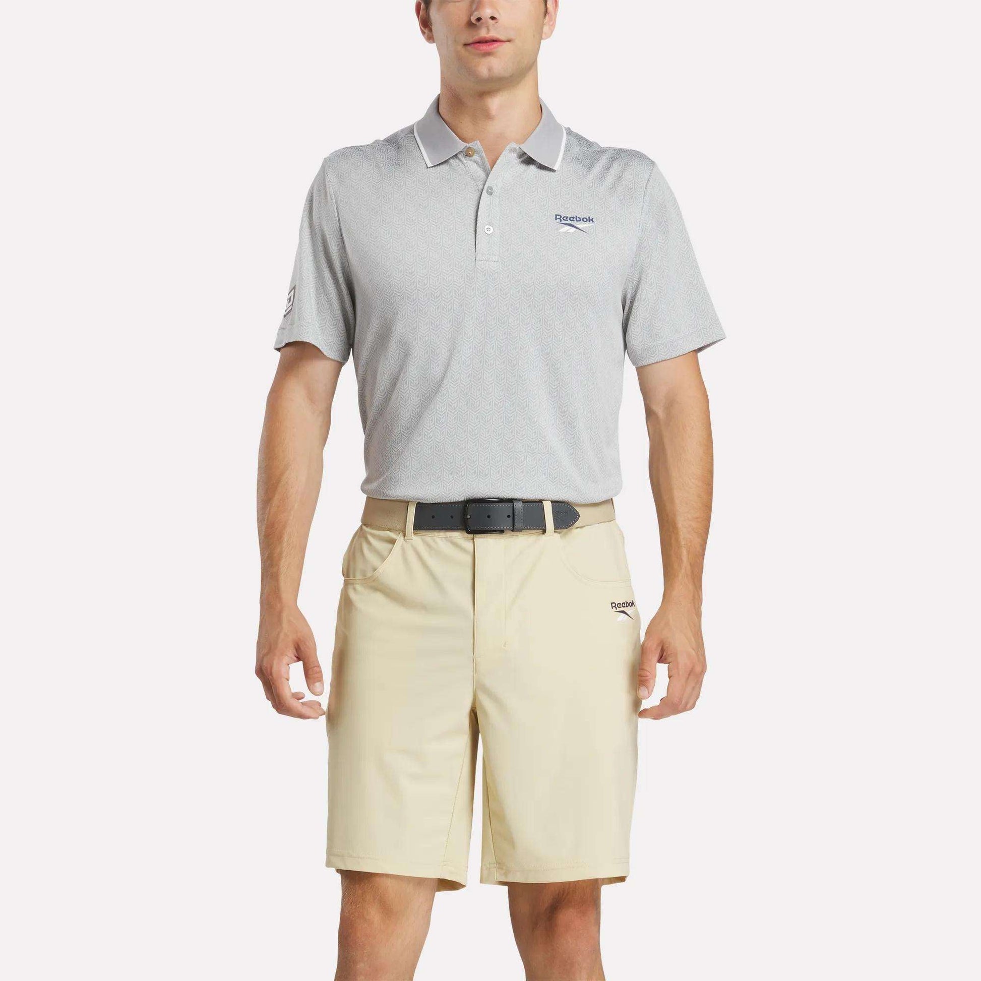 A man wears the Reebok Men's BDC Signature Polo Shirt in light gray, made with UPF-rated RBK-DRY fabric, tucked into beige shorts with a black belt, standing against a plain white background.