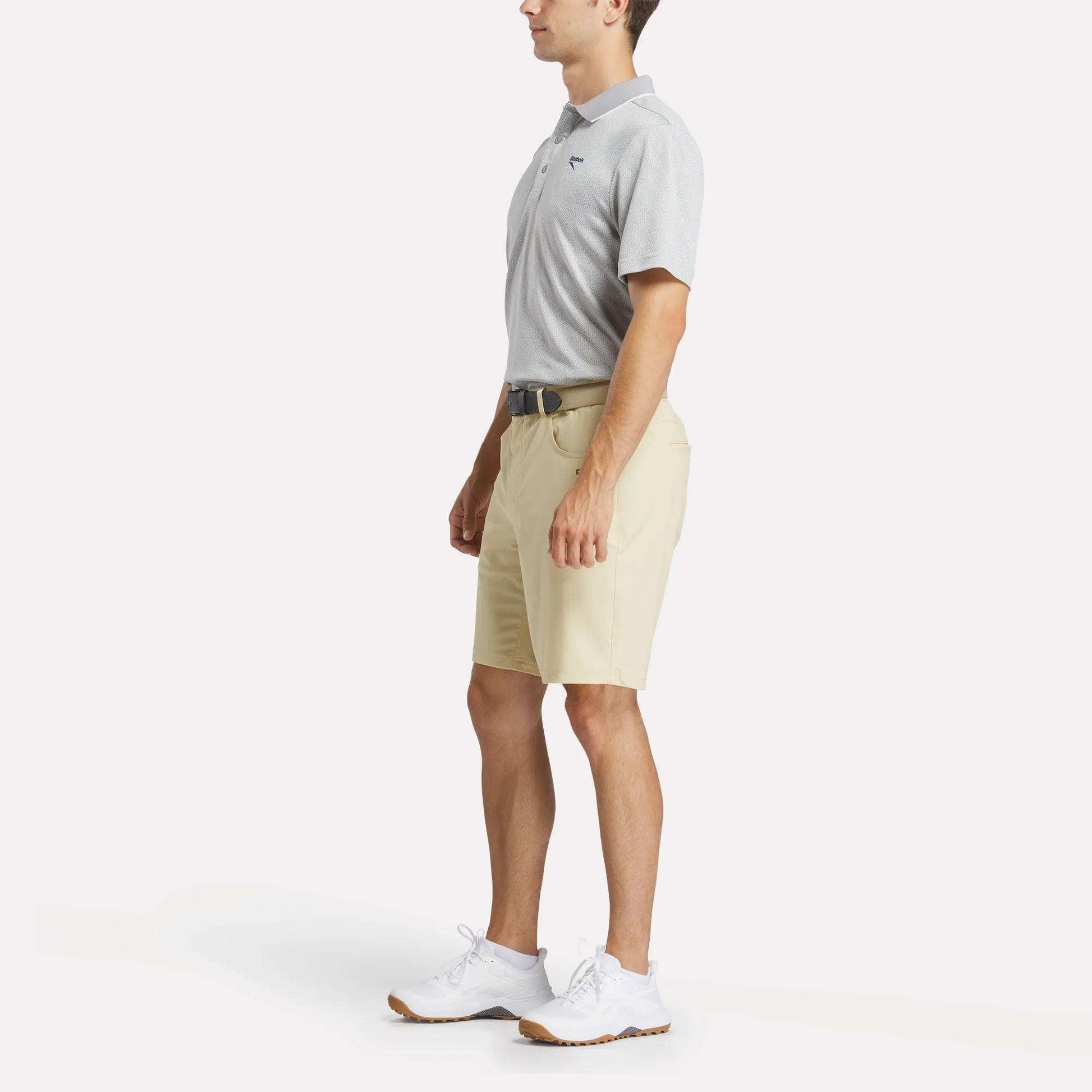 A man in profile wears a light gray Reebok Men's BDC Signature Polo Shirt, beige shorts, white sneakers, and a black belt against a plain white background.