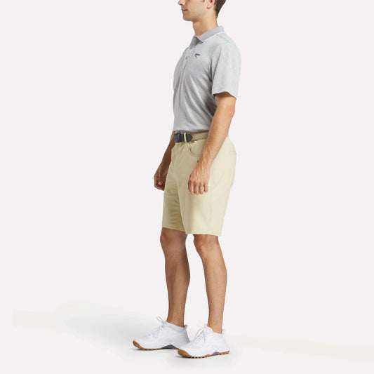 A man in profile wears a light gray Reebok Men's BDC Signature Polo Shirt, beige shorts, white sneakers, and a black belt against a plain white background.