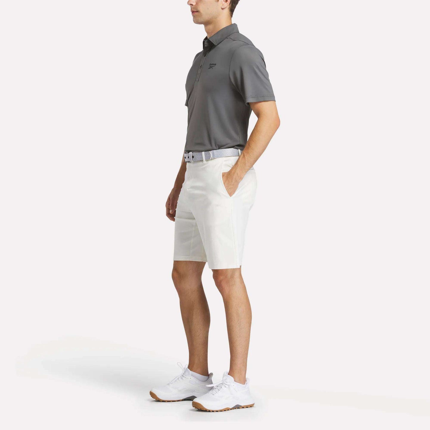 A man stands with hands in his pockets, wearing a gray Reebok Men's BDC Colorblocked Polo Shirt, white shorts, a belt, and white athletic shoes against a plain white background.