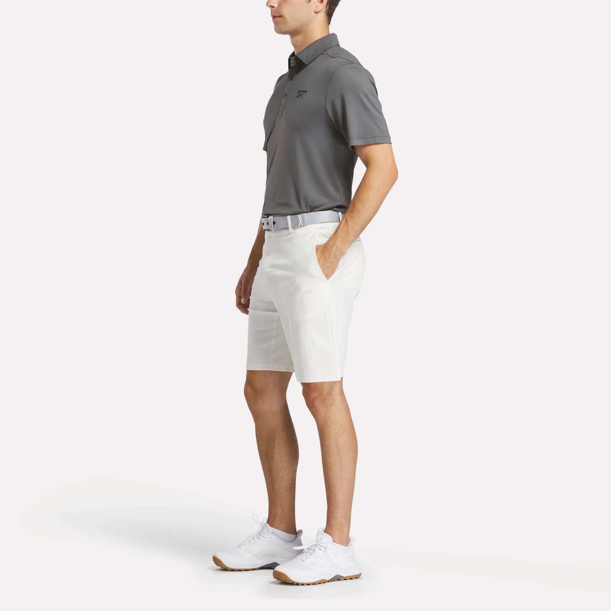 A man stands with hands in his pockets, wearing a gray Reebok Men's BDC Colorblocked Polo Shirt, white shorts, a belt, and white athletic shoes against a plain white background.
