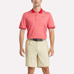 A man faces forward wearing a pink Reebok Men's Country Club Oxford Pique Polo Shirt with white and red trim on the collar and sleeves, an embroidered logo on the left chest, beige shorts, and a black belt.