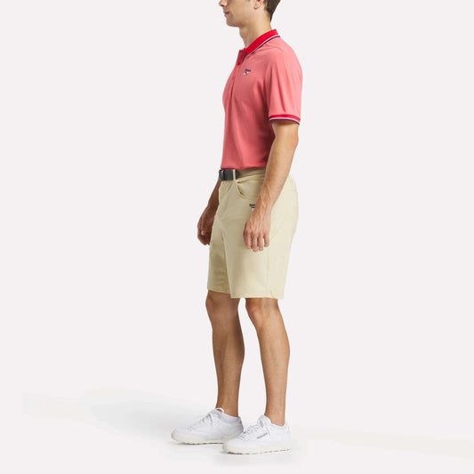 A man stands in profile against a plain white background wearing a red Men's Country Club Oxford Pique Polo Shirt by Reebok, beige shorts, a black belt, and white sneakers.