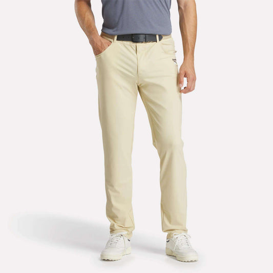 A man stands with his hands in his pockets, wearing light beige Reebok Men's Favorite 5-Pocket Relaxed Fit Golf Pants featuring RBK-DRY tech, a black belt, gray polo, and white sneakers. The image is cropped at the shoulders; his face is not visible.