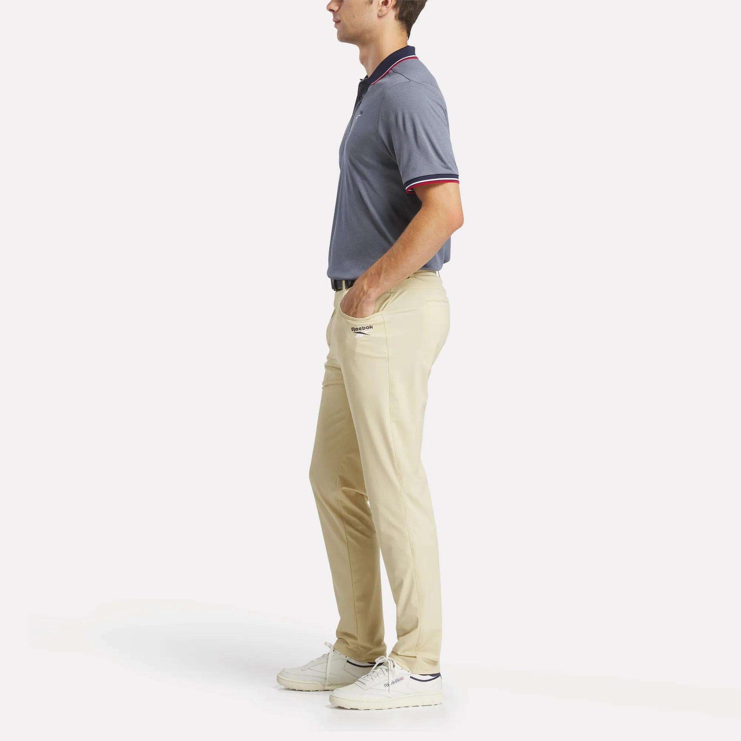 A man stands in profile wearing a short-sleeved blue polo shirt, men's Reebok Favorite 5-Pocket Relaxed Fit Golf Pants, and white sneakers against a plain white background.