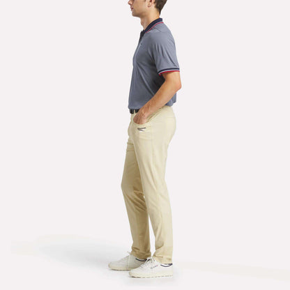 A man stands in profile wearing a short-sleeved blue polo shirt, men's Reebok Favorite 5-Pocket Relaxed Fit Golf Pants, and white sneakers against a plain white background.