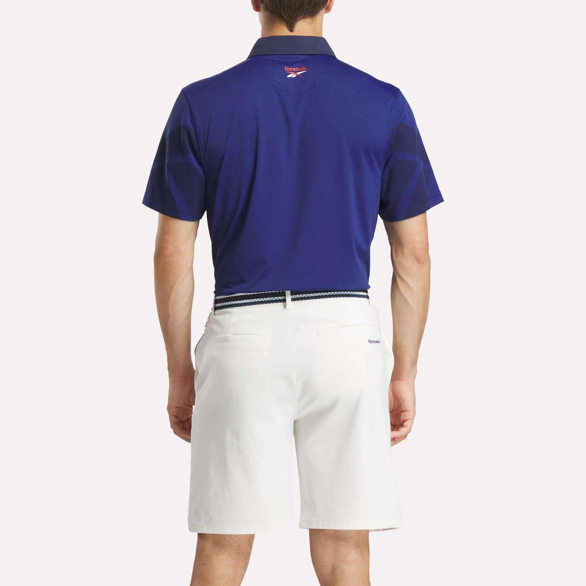 A man faces away, wearing the Reebok Men's Signature Vector Colorblocked Polo Shirt in dark blue, tucked into white shorts with a belt. The background is plain and light-colored.