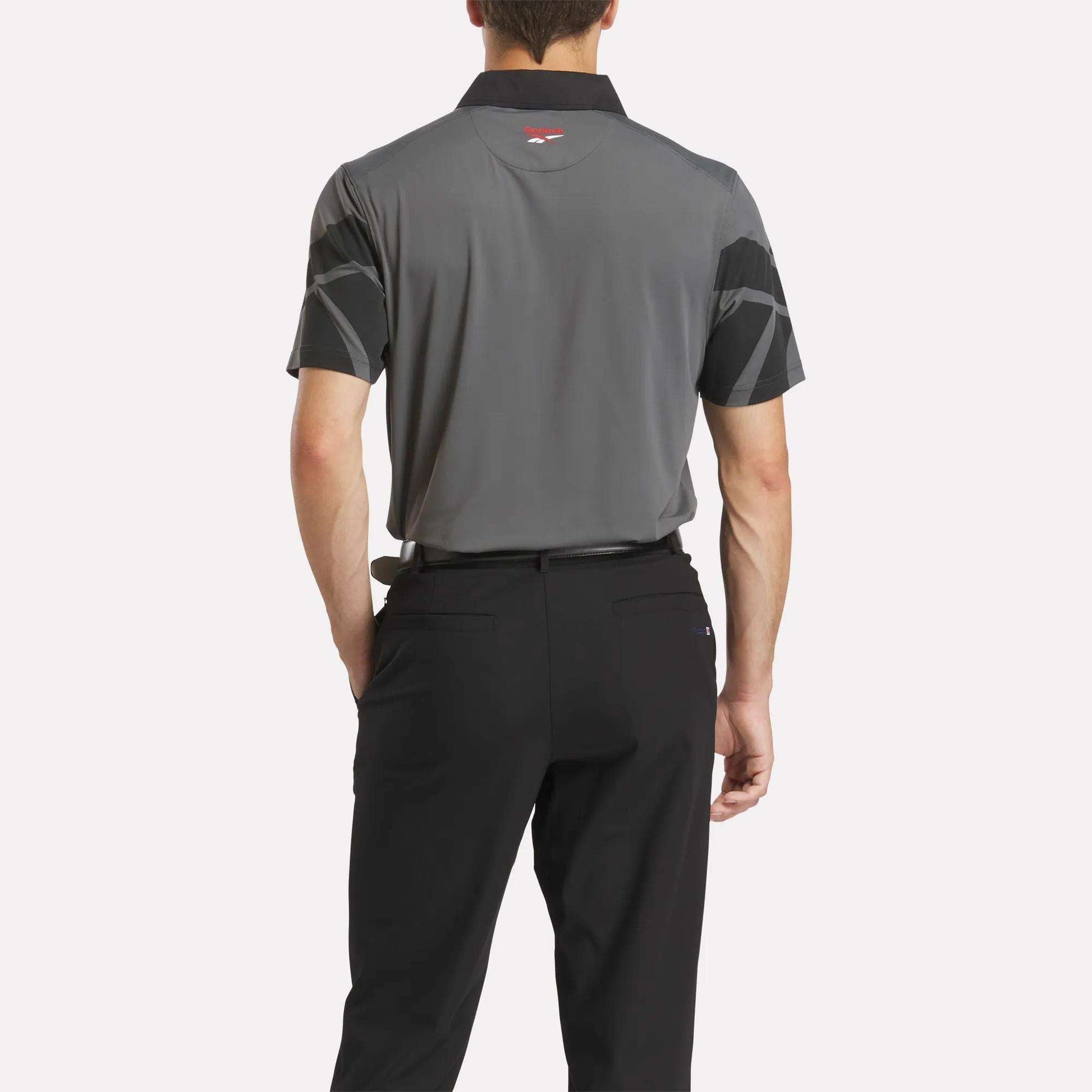 A man with his back to the camera wears a Reebok Men's Signature Vector Colorblocked Polo Shirt in gray and black, paired with black pants, set against a plain white background.