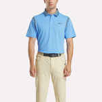 A man stands facing forward in a light blue Reebok Men's Varsity Polo Shirt and beige pants with a black belt against a plain white background.