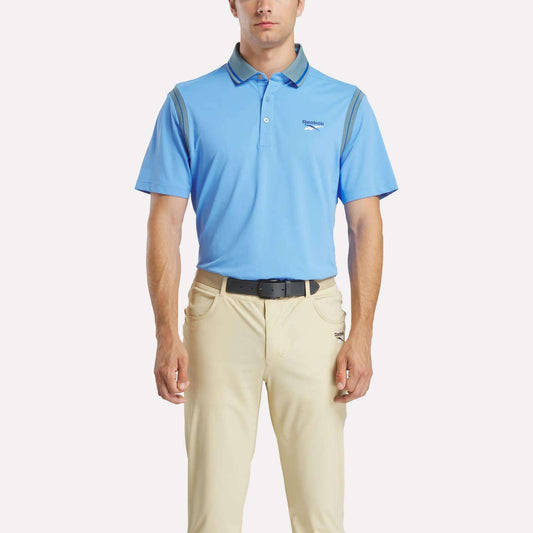 A man stands facing forward in a light blue Reebok Men's Varsity Polo Shirt and beige pants with a black belt against a plain white background.