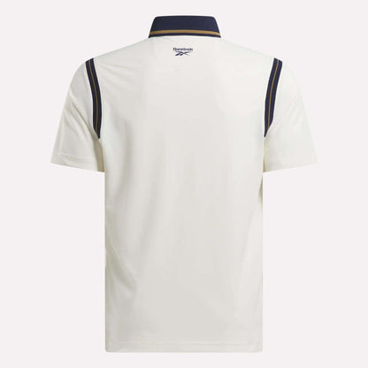 Back view of the Men's Varsity Polo Shirt by Reebok, crafted with RBK-DRY fabric. Features a white short-sleeve design, navy collar, navy shoulder stripes, and a Reebok logo at the upper back below the collar.