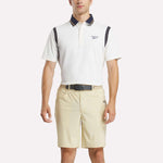 A man stands facing forward with hands relaxed, wearing a white Reebok Men's Varsity Polo Shirt with dark accents and beige shorts against a plain light background.