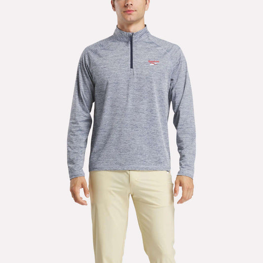 A man stands facing forward, arms at his sides, wearing a Reebok Men's Cationic Wicking Quarter-Zip Sweatshirt in light blue and beige pants against a plain white background.