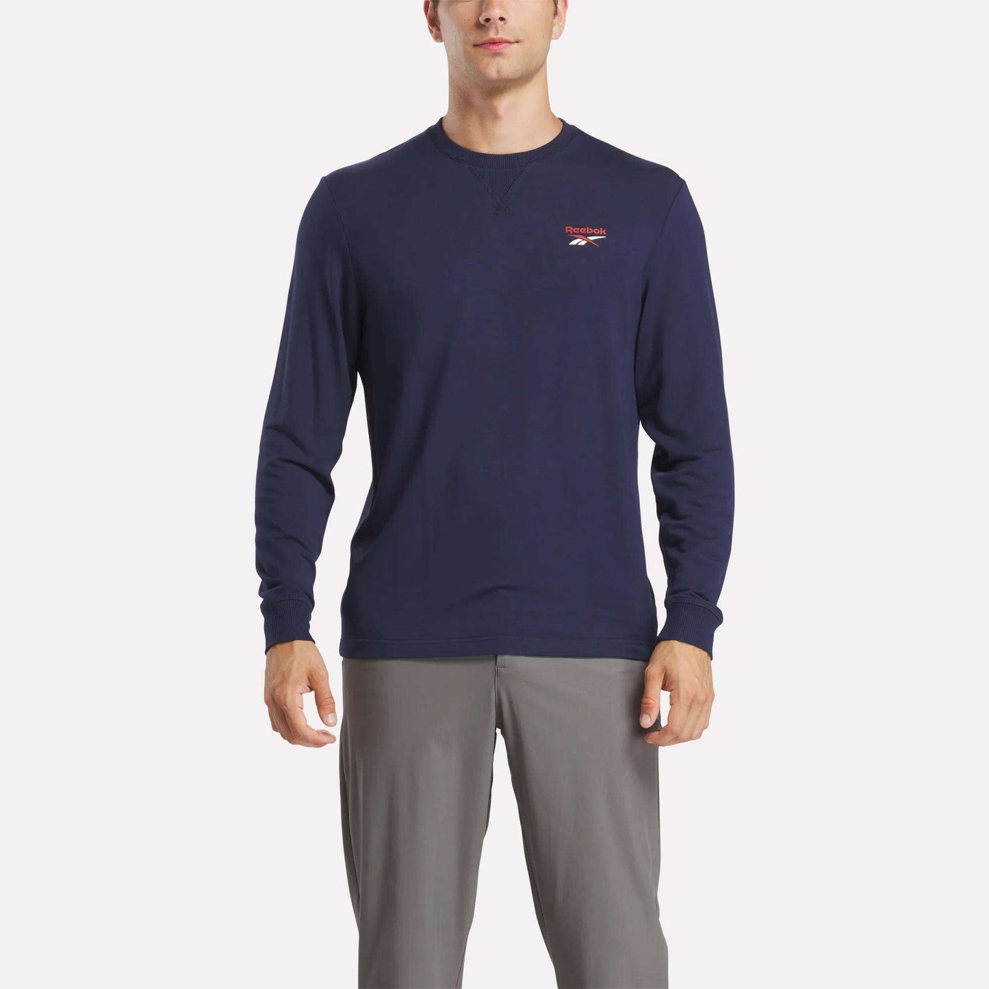 A man wears a Reebok Men's Long Sleeve Golf Sweatshirt in navy blue with UPF 30+ and a small red and white chest logo, paired with light gray pants, standing against a white background.