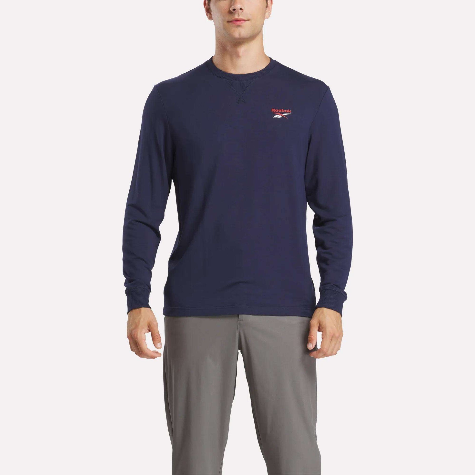 A man wears a Reebok Men's Long Sleeve Golf Sweatshirt in navy blue with UPF 30+ and a small red and white chest logo, paired with light gray pants, standing against a white background.