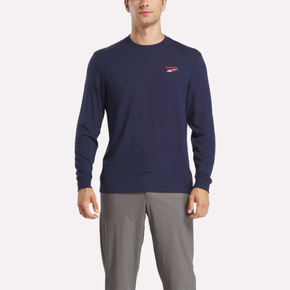 A man wears a Reebok Men's Long Sleeve Golf Sweatshirt in navy blue with UPF 30+ and a small red and white chest logo, paired with light gray pants, standing against a white background.