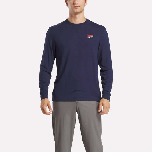 A man wears a Reebok Men's Long Sleeve Golf Sweatshirt in navy blue with UPF 30+ and a small red and white chest logo, paired with light gray pants, standing against a white background.