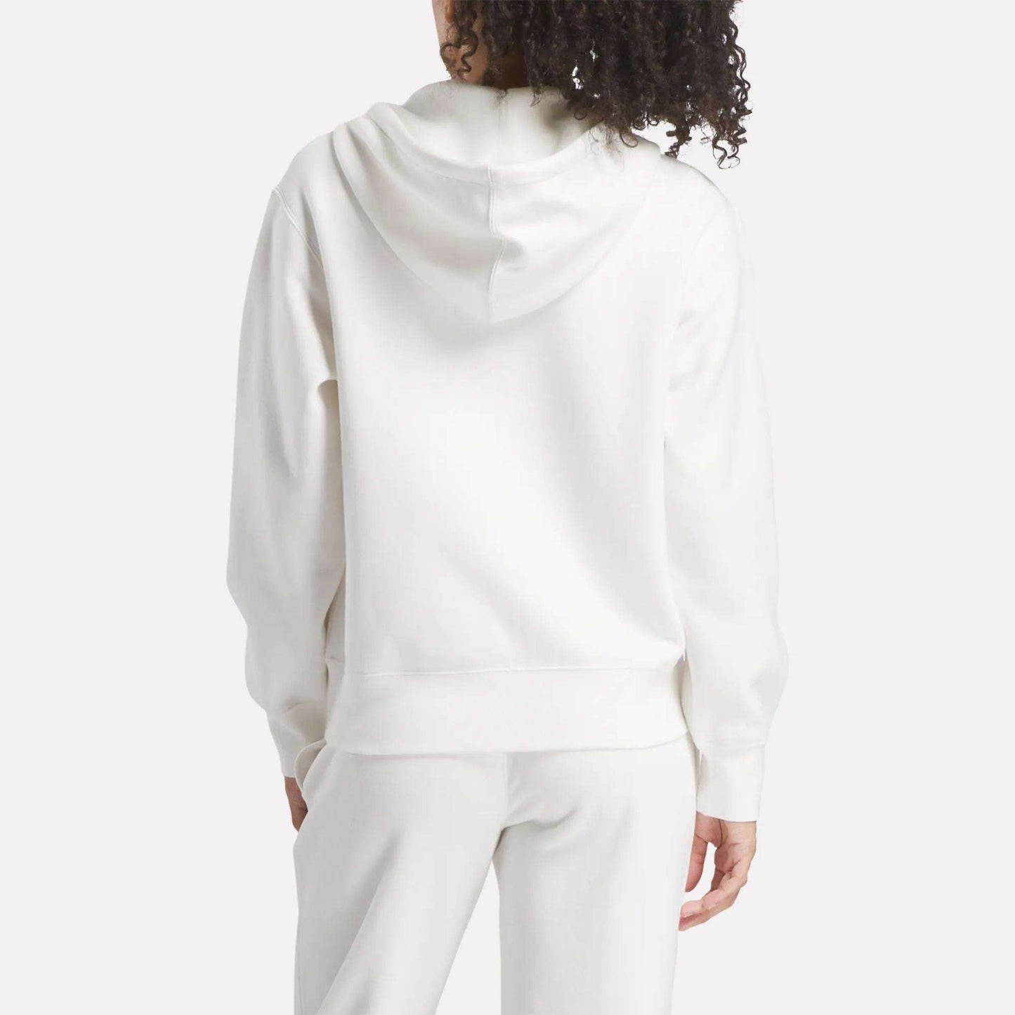 A person with curly hair, seen from behind, wears a Reebok Women's Small Vector Full Zip Sweatshirt in white and matching white pants. The hoodie is down, set against a plain light-colored background.