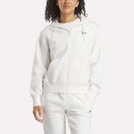 A woman wears the Reebok Women's Small Vector Full Zip Sweatshirt in white, paired with matching sweatpants, standing against a plain light background with one hand in her pocket and a relaxed expression.