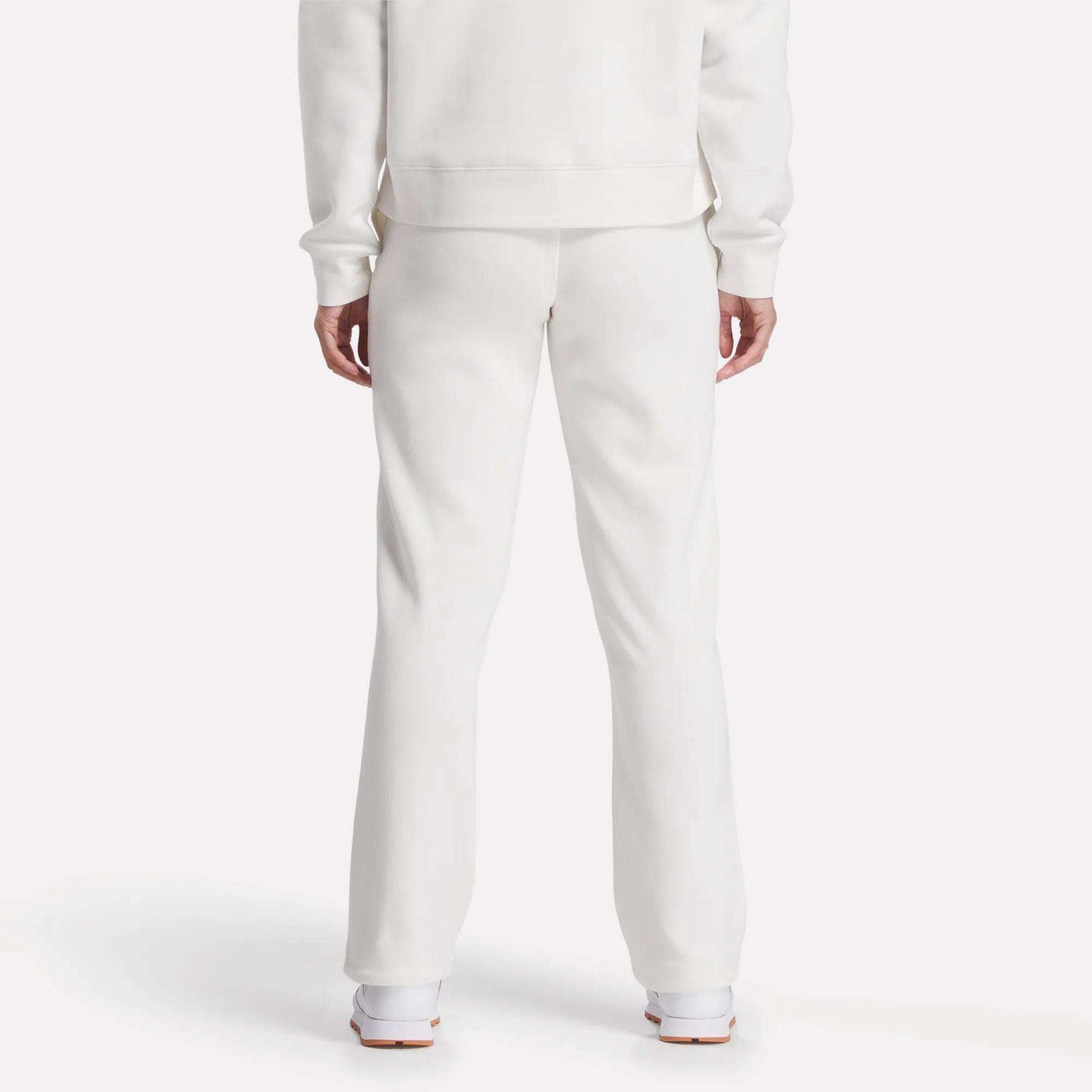 A person is seen from behind, showing only lower back and legs, wearing Reebok Women's Cozy Classic Sweatpants in soft white cotton blend with a matching long-sleeve top and white shoes, set against a plain white background.