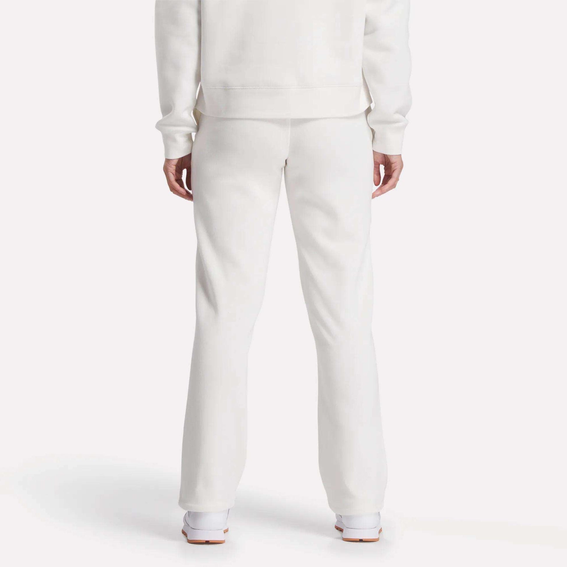 A person is seen from behind, showing only lower back and legs, wearing Reebok Women's Cozy Classic Sweatpants in soft white cotton blend with a matching long-sleeve top and white shoes, set against a plain white background.