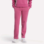 Visible from the shoulders down, a person models Reebok Women's Cozy Classic Sweatpants in bright pink with a matching sweatshirt and white sneakers, standing hands-in-pockets against a plain white background.