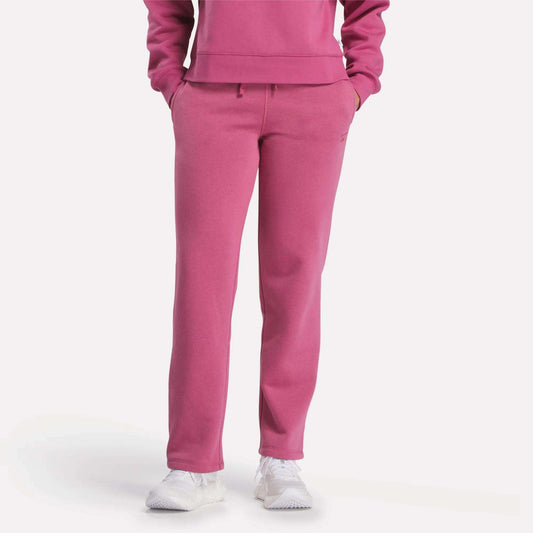 Visible from the shoulders down, a person models Reebok Women's Cozy Classic Sweatpants in bright pink with a matching sweatshirt and white sneakers, standing hands-in-pockets against a plain white background.