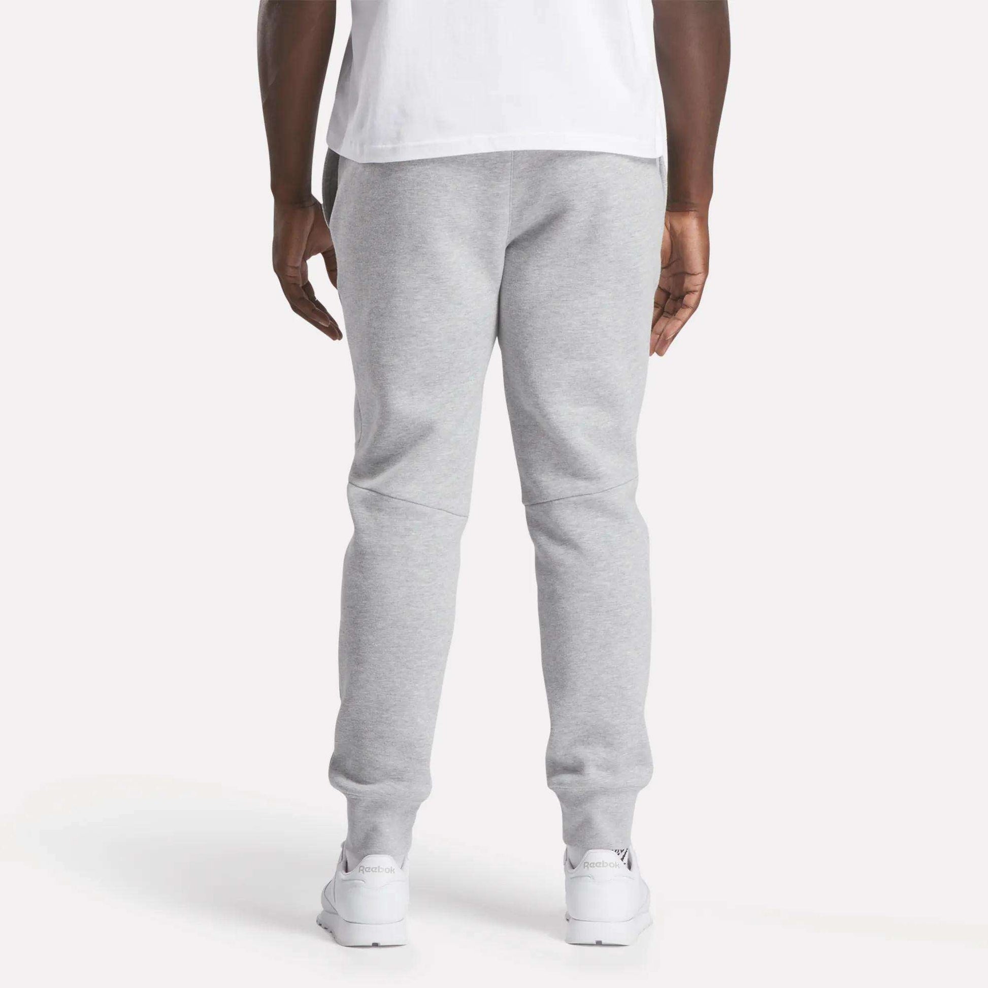 A person stands facing away from the camera, wearing a white T-shirt, Reebok Men's Motion Joggers in light gray slim fit, and white sneakers against a plain light background.