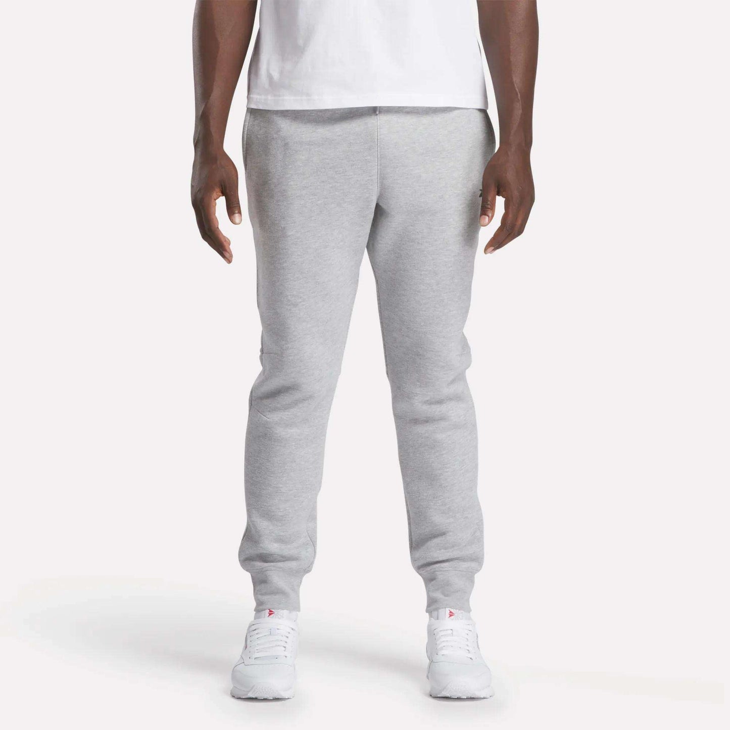 A person stands facing forward against a plain white background, wearing a white t-shirt, Reebok Men's Motion Joggers in a slim fit, and white sneakers.