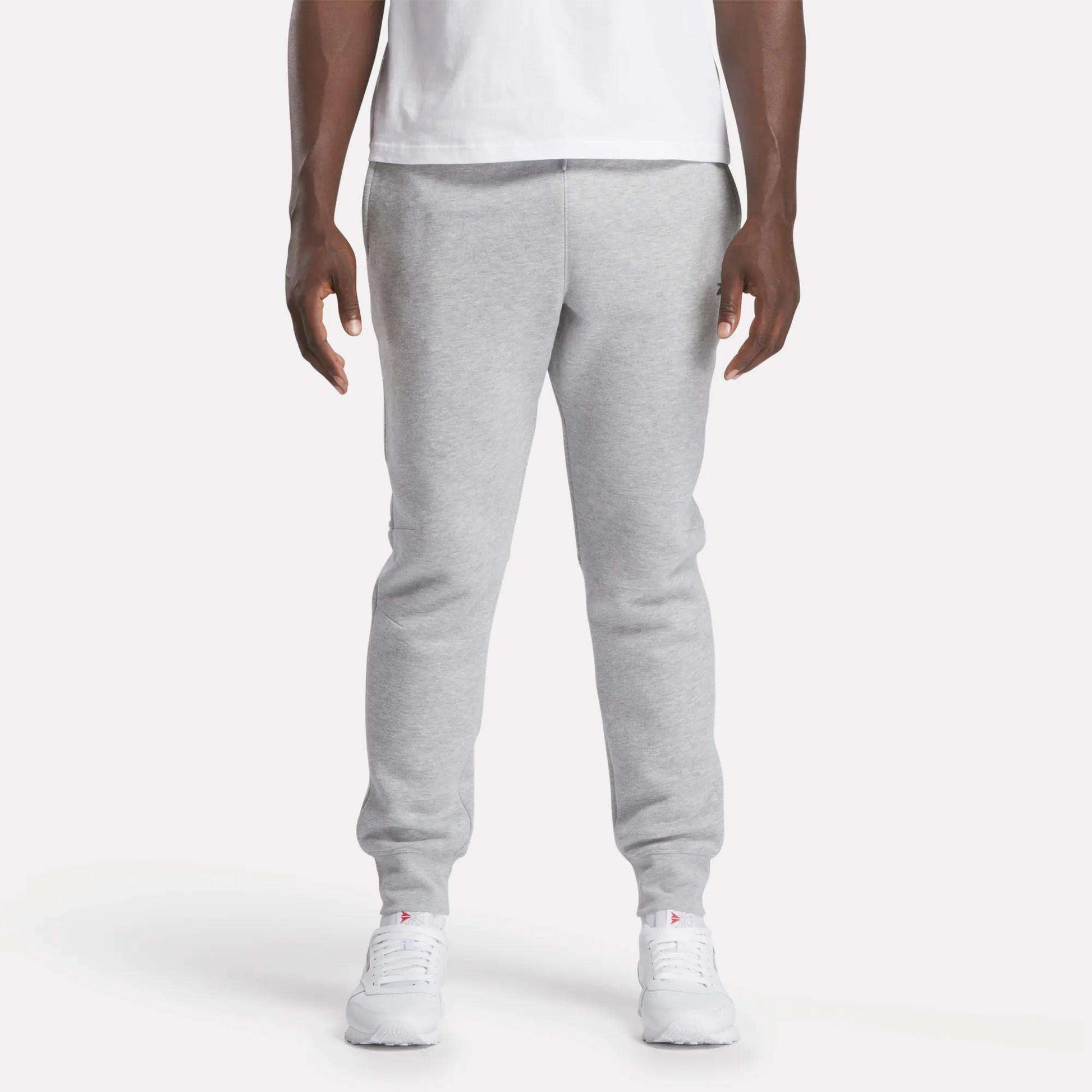 A person stands facing forward against a plain white background, wearing a white t-shirt, Reebok Men's Motion Joggers in a slim fit, and white sneakers.