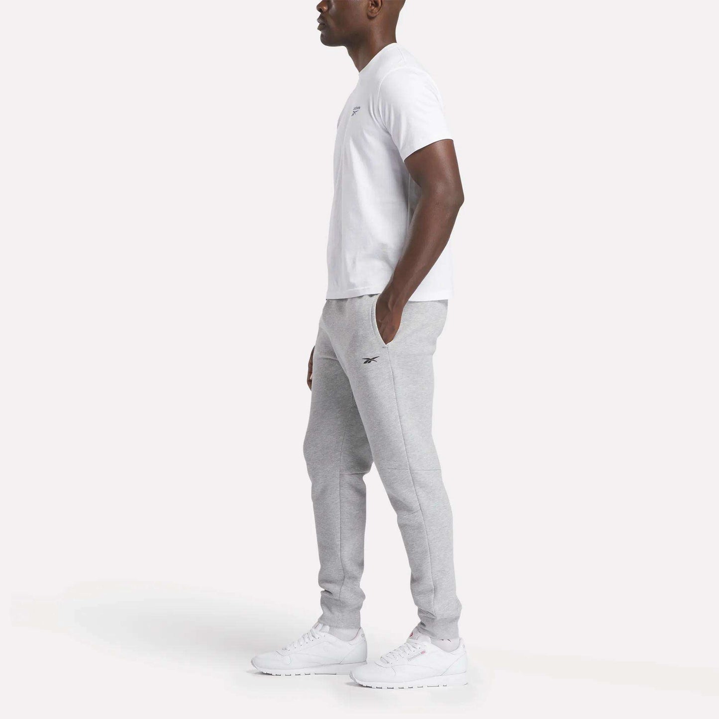A man stands in profile against a plain white background wearing Reebok Men's Motion Joggers, a white T-shirt, and white athletic shoes.