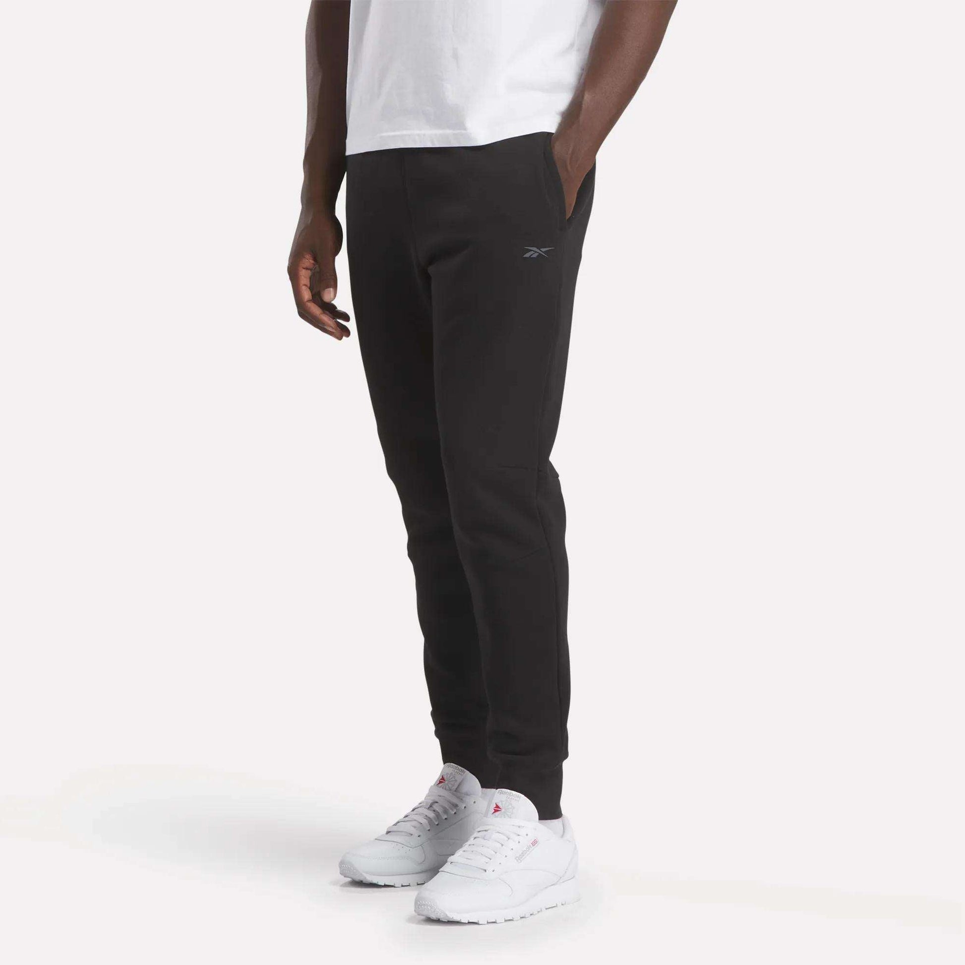 A person wearing a white T-shirt, Reebok Men's Motion Joggers with hands in pockets, and white athletic shoes stands against a plain, light background.