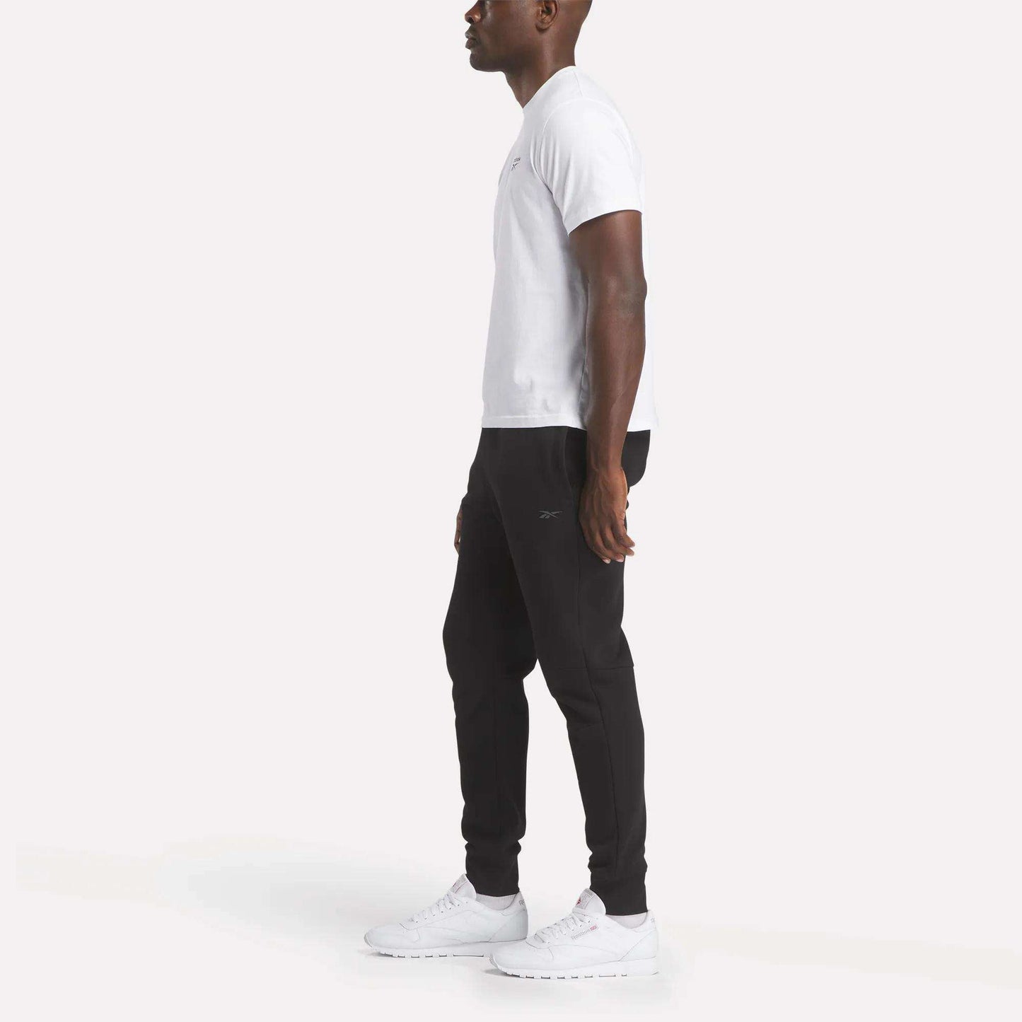 A man stands in profile wearing Reebok Men's Motion Joggers, a white t-shirt, and white sneakers against a plain white background.