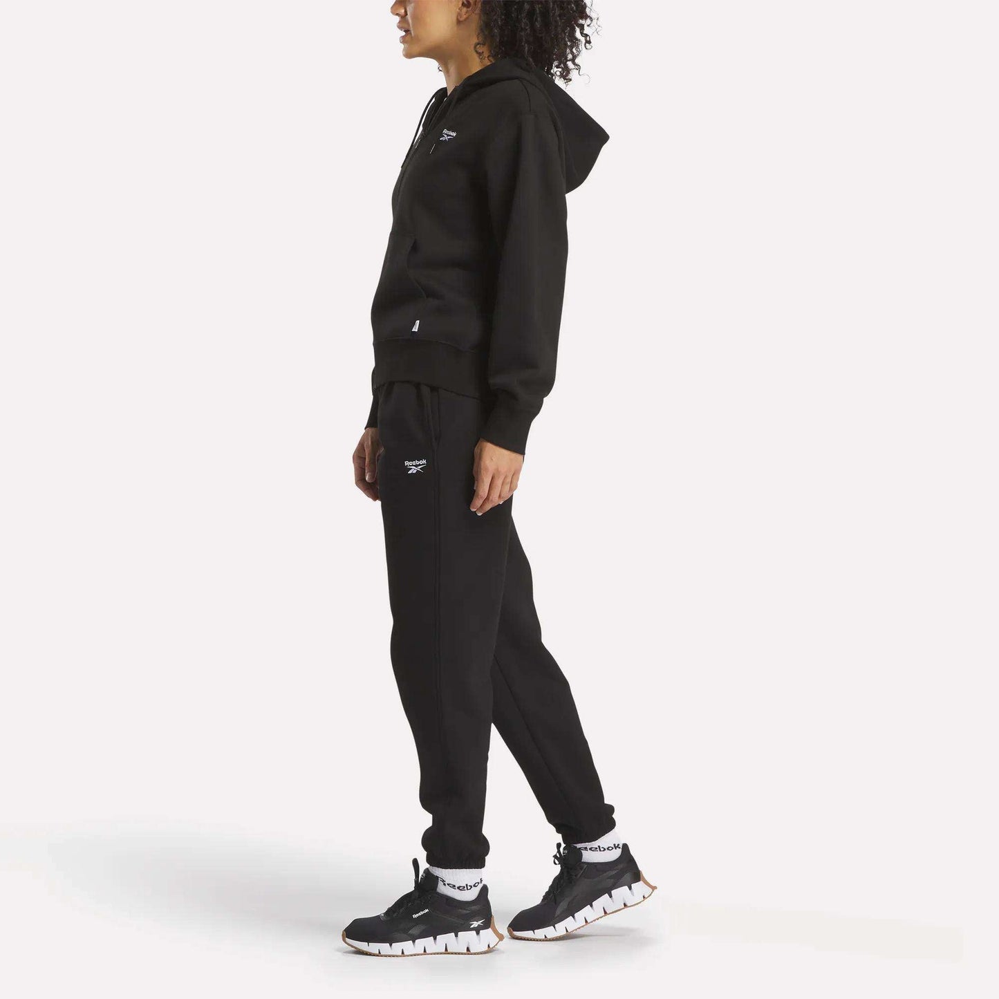 A person in profile wears Reebok Women's Medium Vector Sweatpants featuring a small white logo, paired with a black hooded sweatshirt and black sneakers with white soles, all shown against a plain white background.