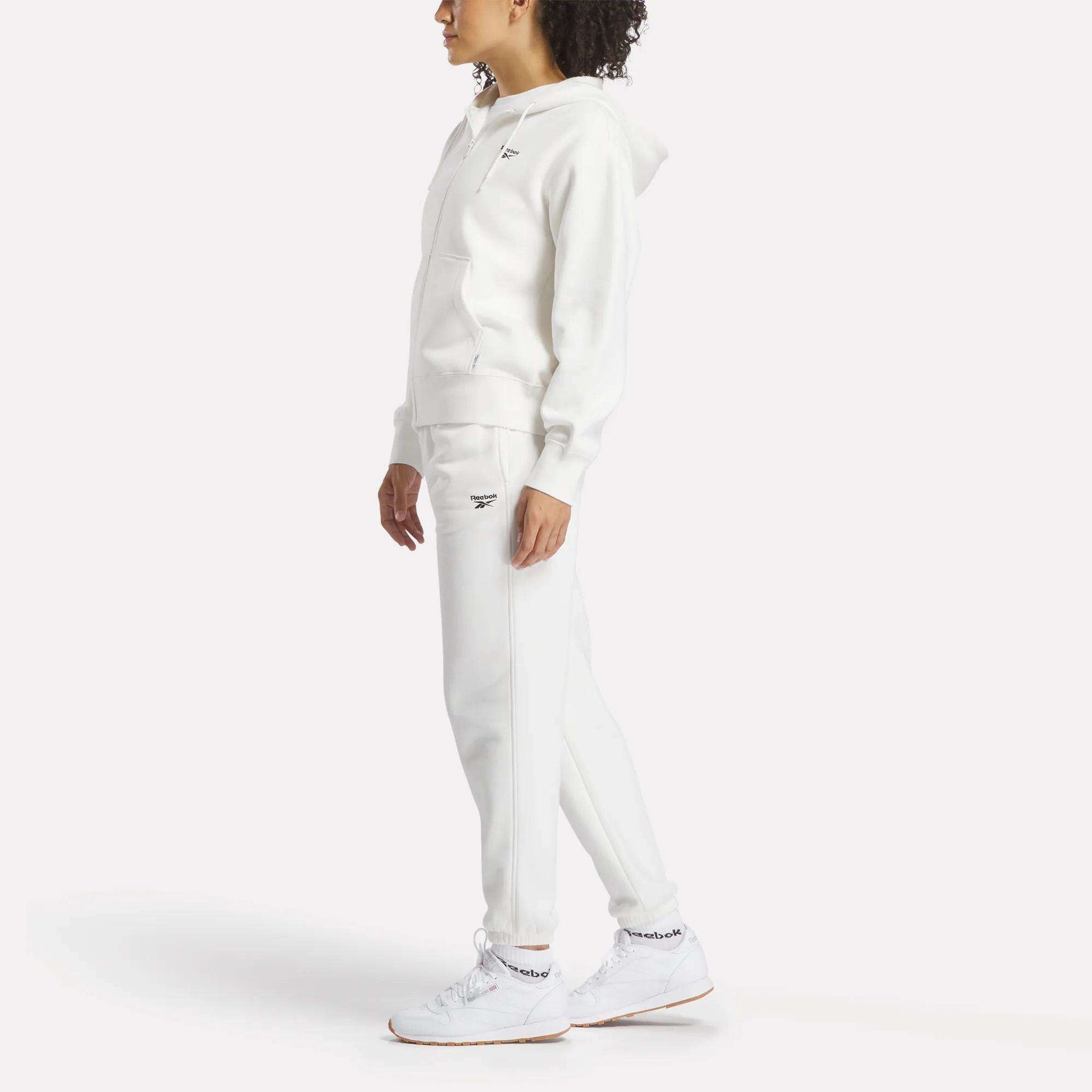 A person stands against a white background wearing an all-white Reebok hoodie, Women's Medium Vector Sweatpants with a small logo, and sneakers. The cotton-blend sweatpants are by Reebok, and the person's face is turned slightly away.