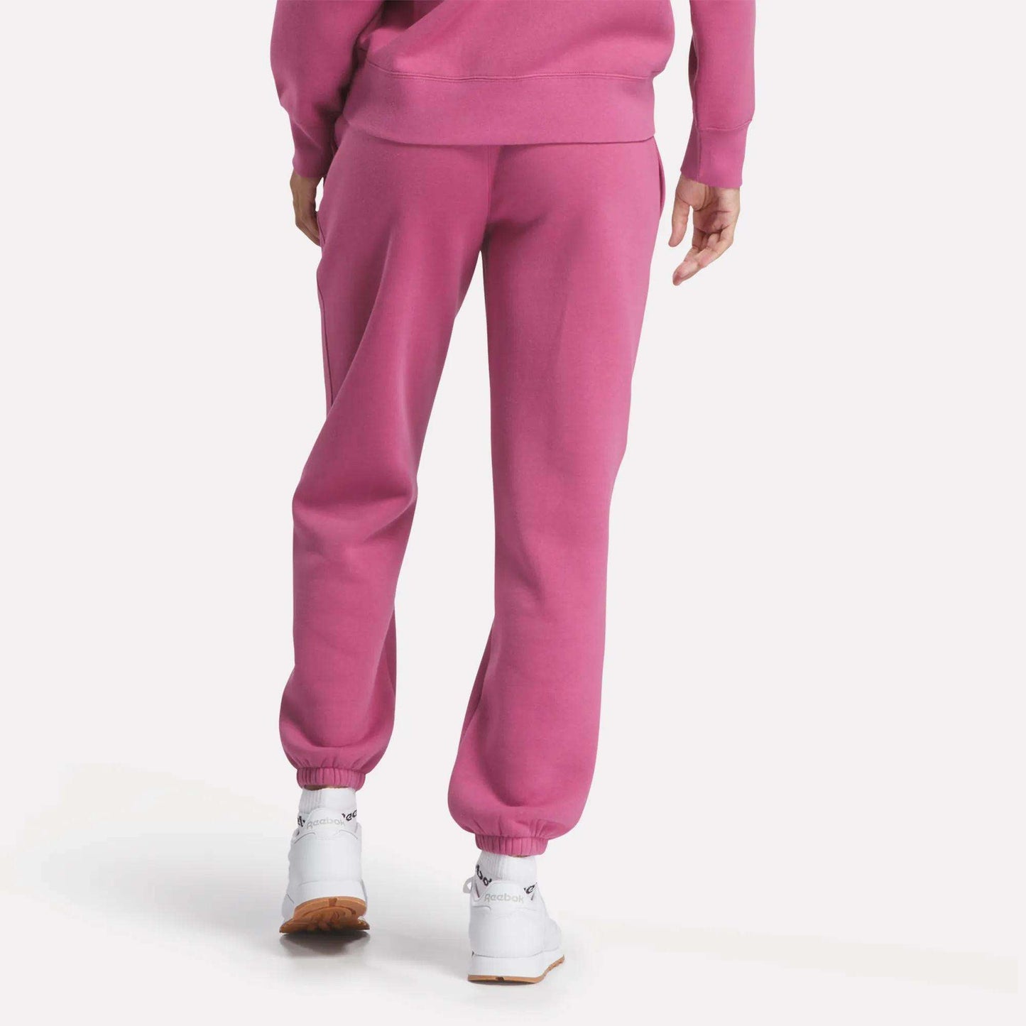 A person faces away in Reebok Women's Medium Vector Sweatpants and a matching sweatshirt, bright pink in color, paired with white sneakers against a plain white background.