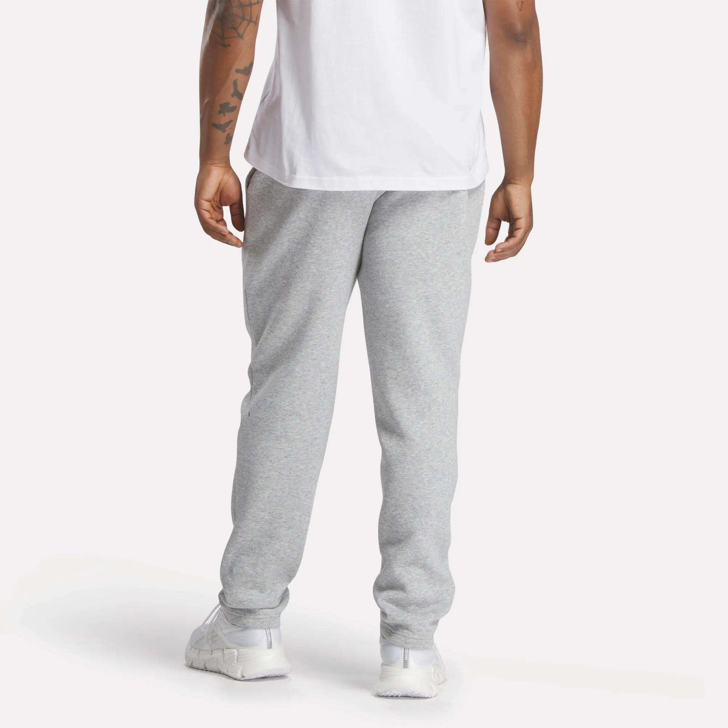 A person is seen from behind, standing against a plain background, wearing a white T-shirt, Reebok Men's Patent Pants (slim fit, adjustable waistband), and white sneakers.