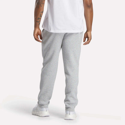A person is seen from behind, standing against a plain background, wearing a white T-shirt, Reebok Men's Patent Pants (slim fit, adjustable waistband), and white sneakers.