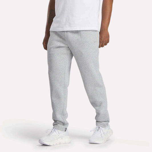 A person wearing Reebok Men's Patent Pants in slim fit light gray with an adjustable waistband is shown from the waist down against a plain background. White t-shirt and sneakers are also visible.