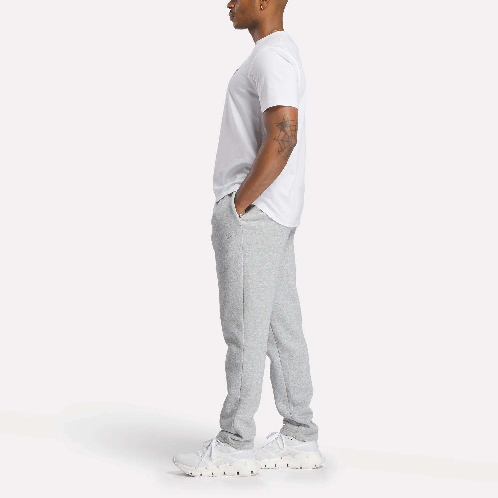A man stands in profile with hands in pockets, wearing a white T-shirt, white sneakers, and slim fit light gray Reebok Men's Patent Pants featuring an adjustable waistband, against a plain white background.